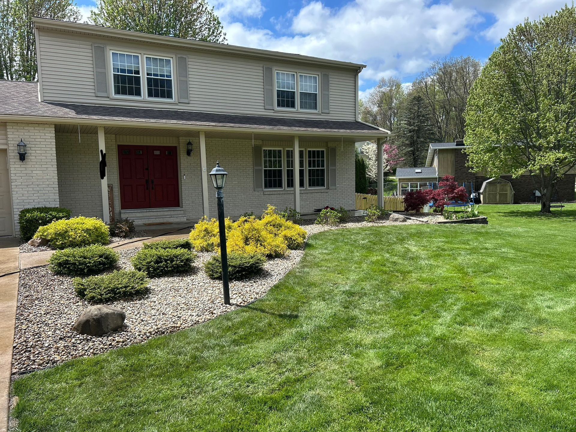 Two-story house with red door, landscaped front yard, yellow bushes, and green grass under a blue sky.
