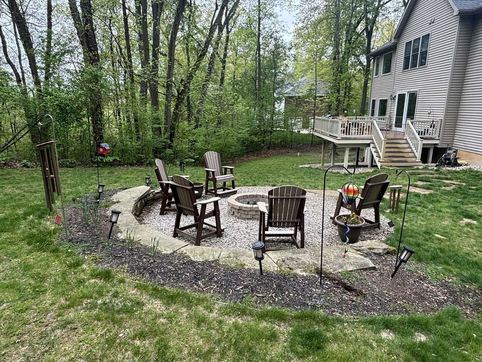 A backyard fire pit with chairs arranged around it, a deck, and trees in the background.
