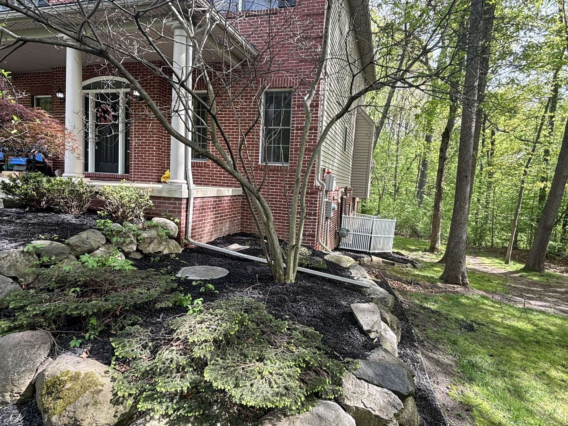 Brick house exterior with landscaping, a tree, and a wooded backdrop.