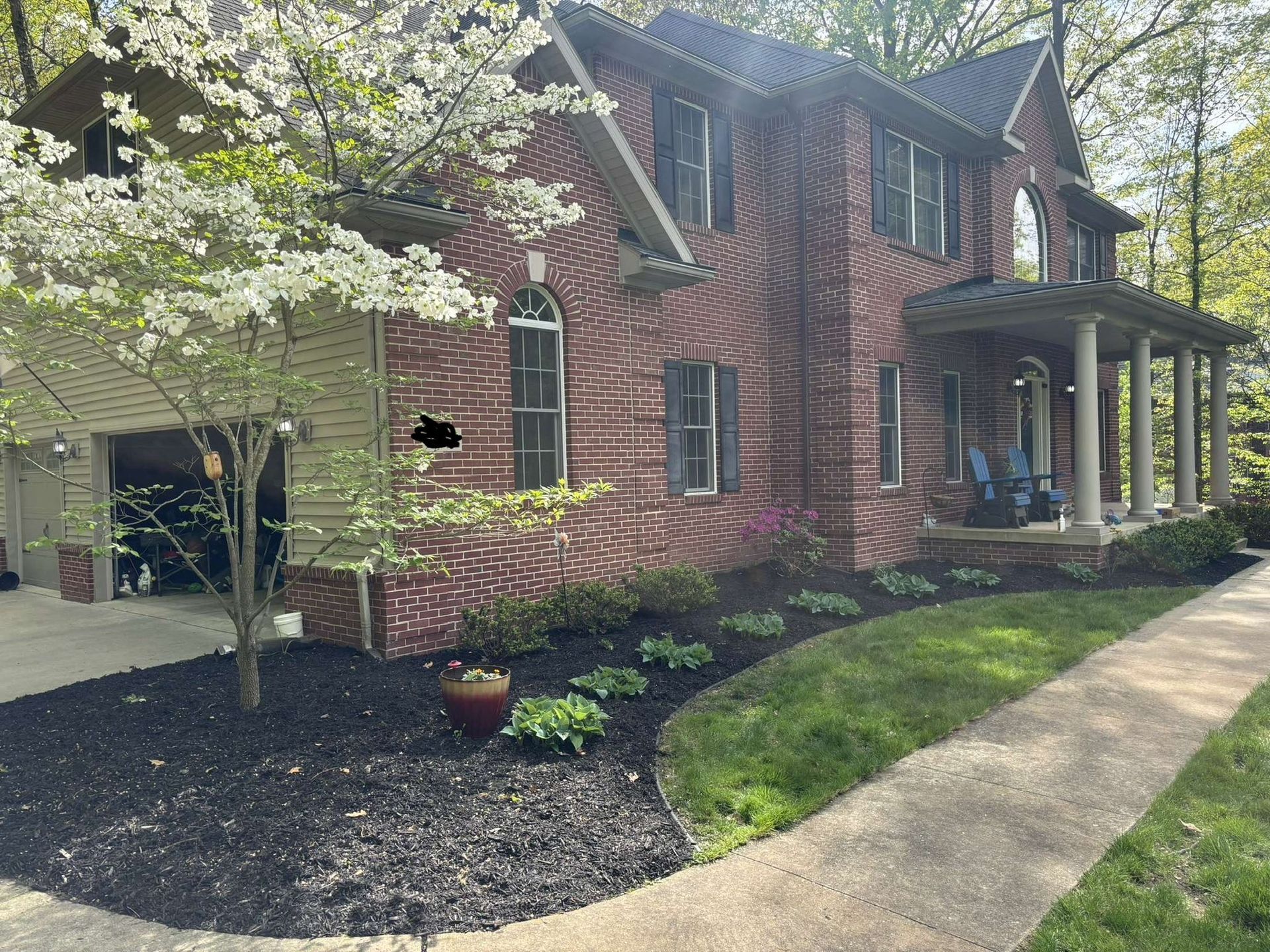 Two-story brick house with porch, garage, and landscaped yard with black mulch and a flowering tree.