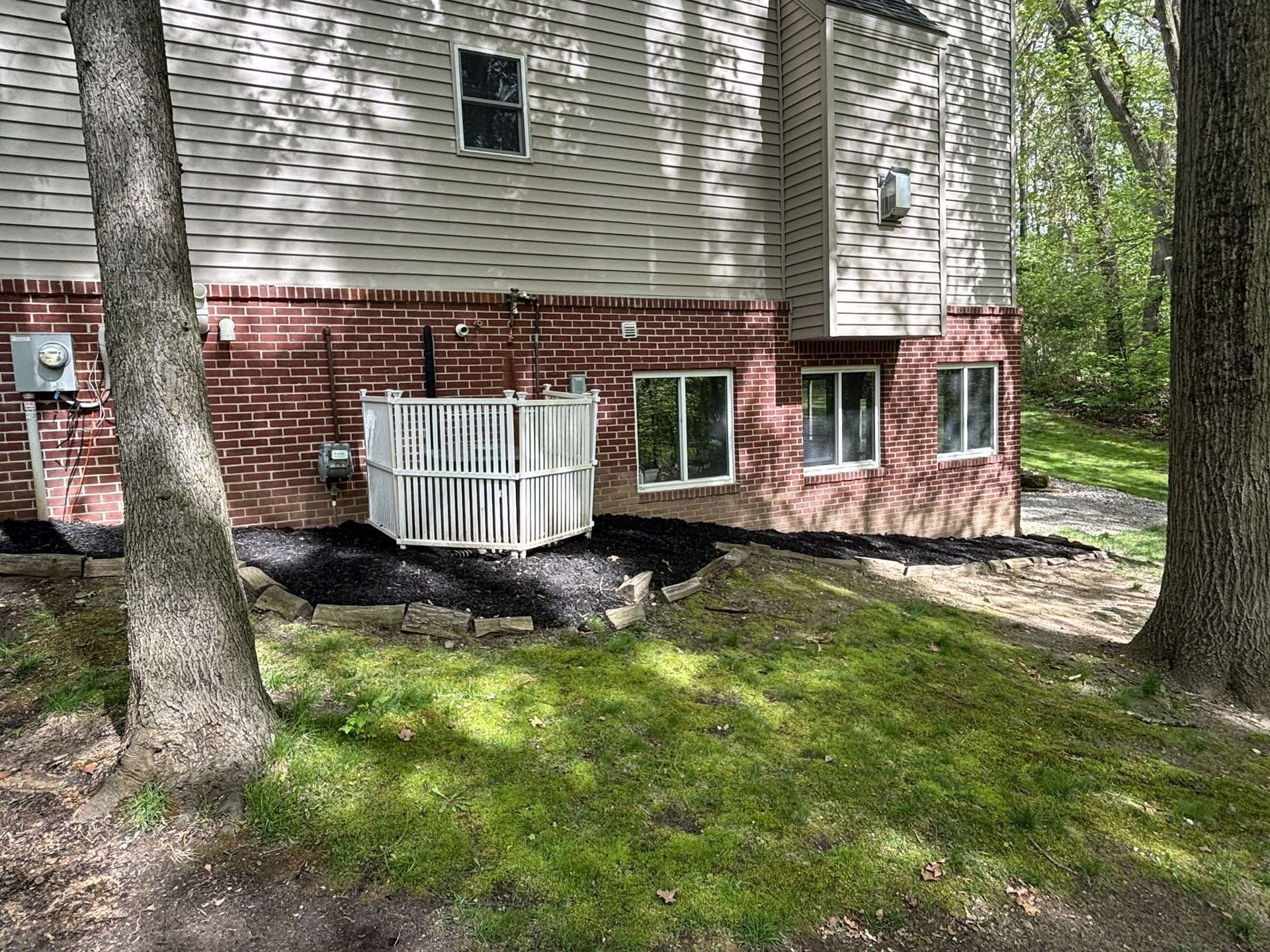 Red brick and beige sided house with a white AC unit and landscaping, surrounded by trees.