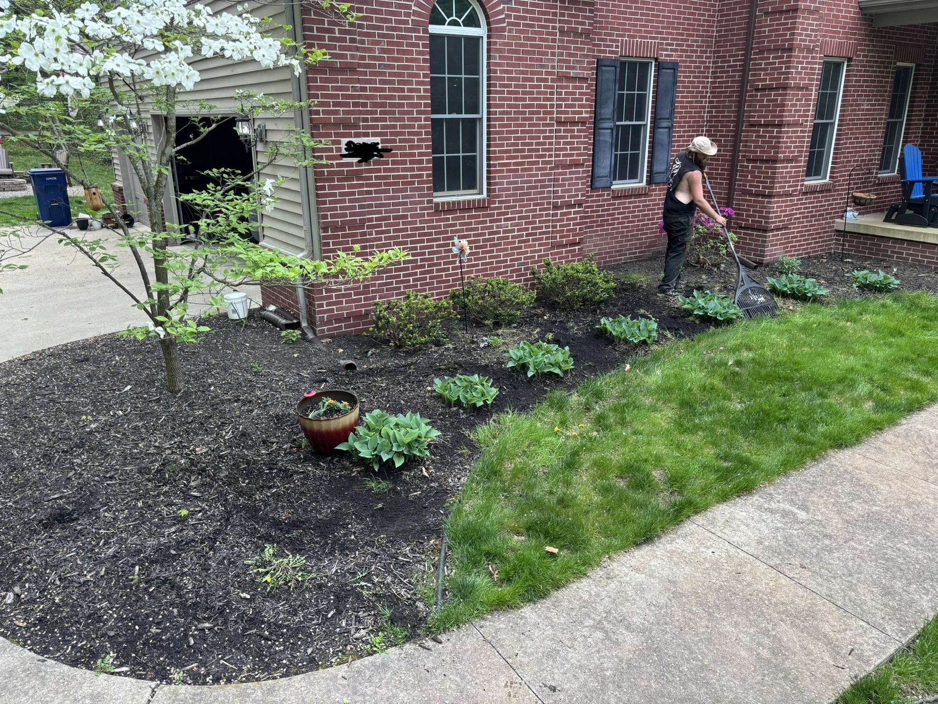 Woman watering plants in a front yard flower bed next to a brick house and sidewalk.