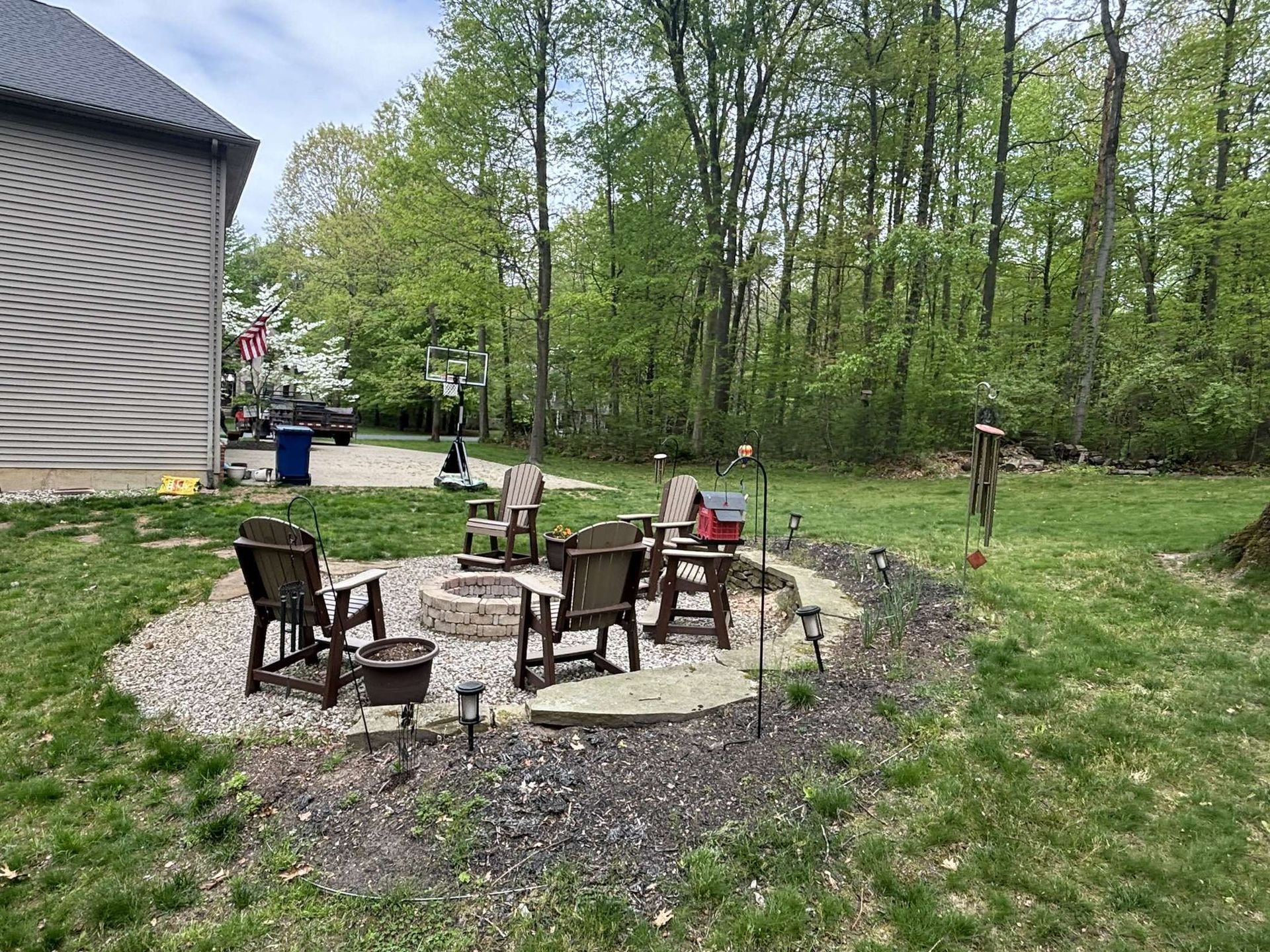Fire pit with six chairs in a backyard, surrounded by gravel and greenery.