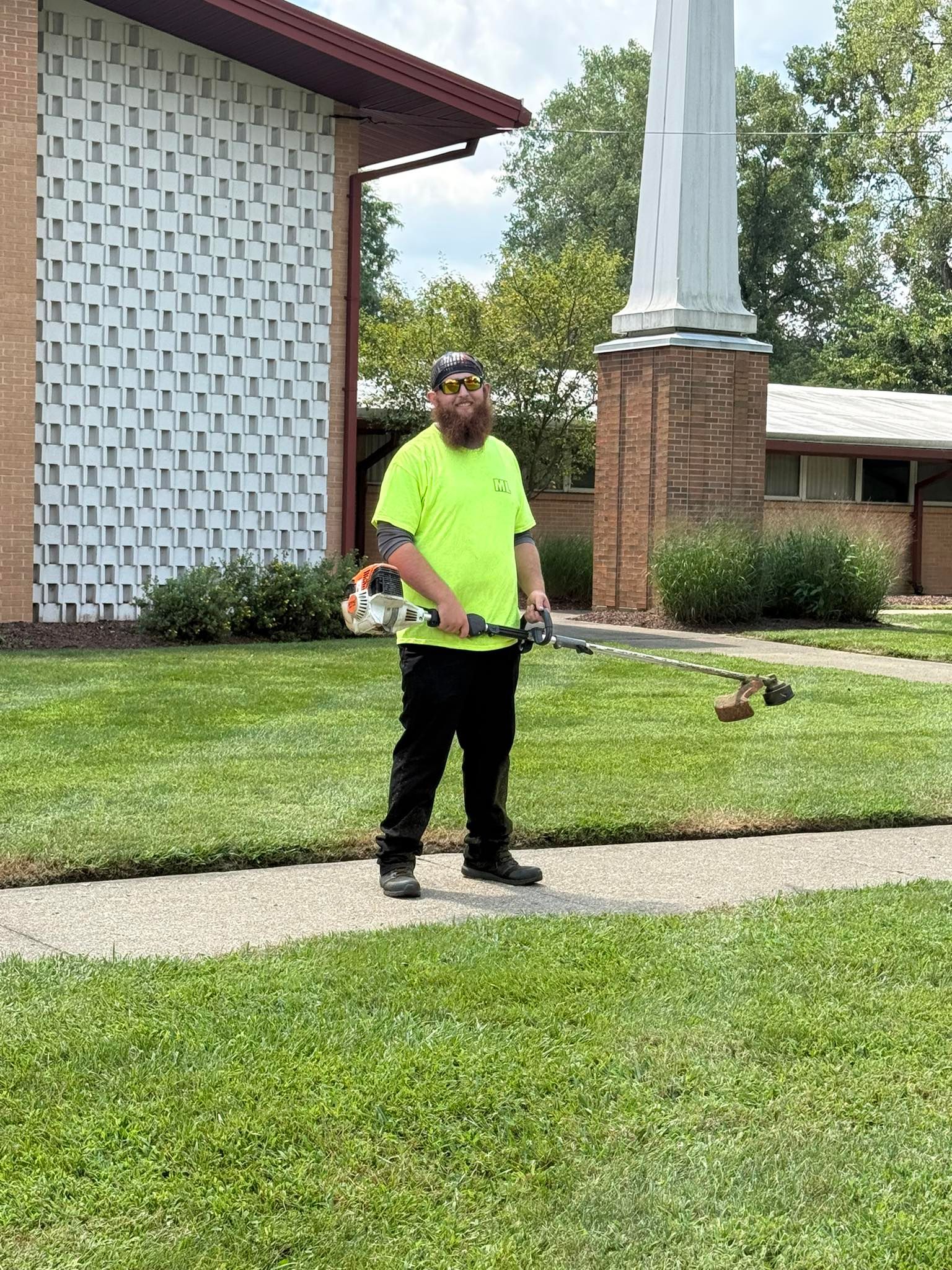 Man in neon green shirt, black pants, and hat uses a weed whacker on a sidewalk.