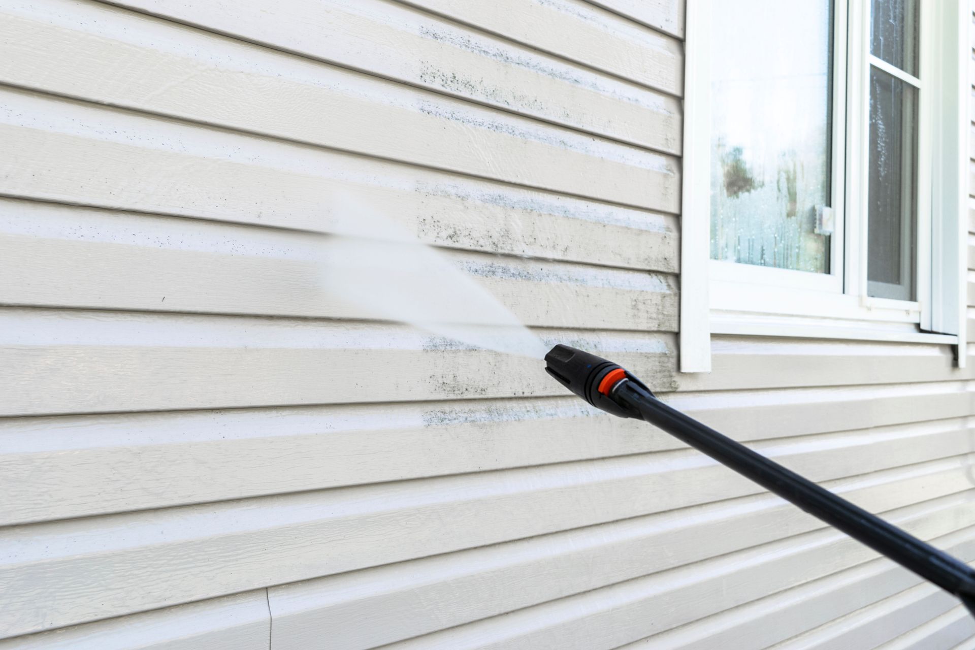 Pressure washing a light-colored house siding, cleaning off mildew and dirt, next to a window.