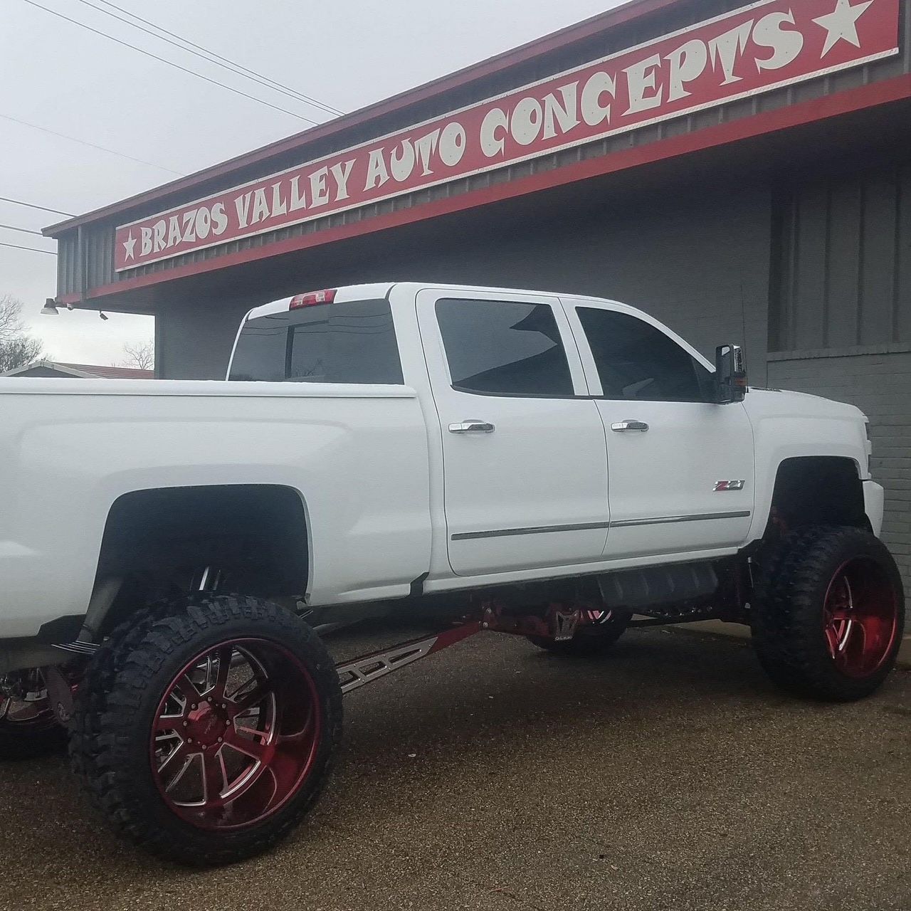 White lifted truck with red rims parked in front of Brazos Valley Auto Concepts shop.