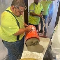 Men in work clothes, one pouring from an orange bucket, inside a construction site.