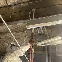 Person in protective suit spraying insulation in a building's ceiling.