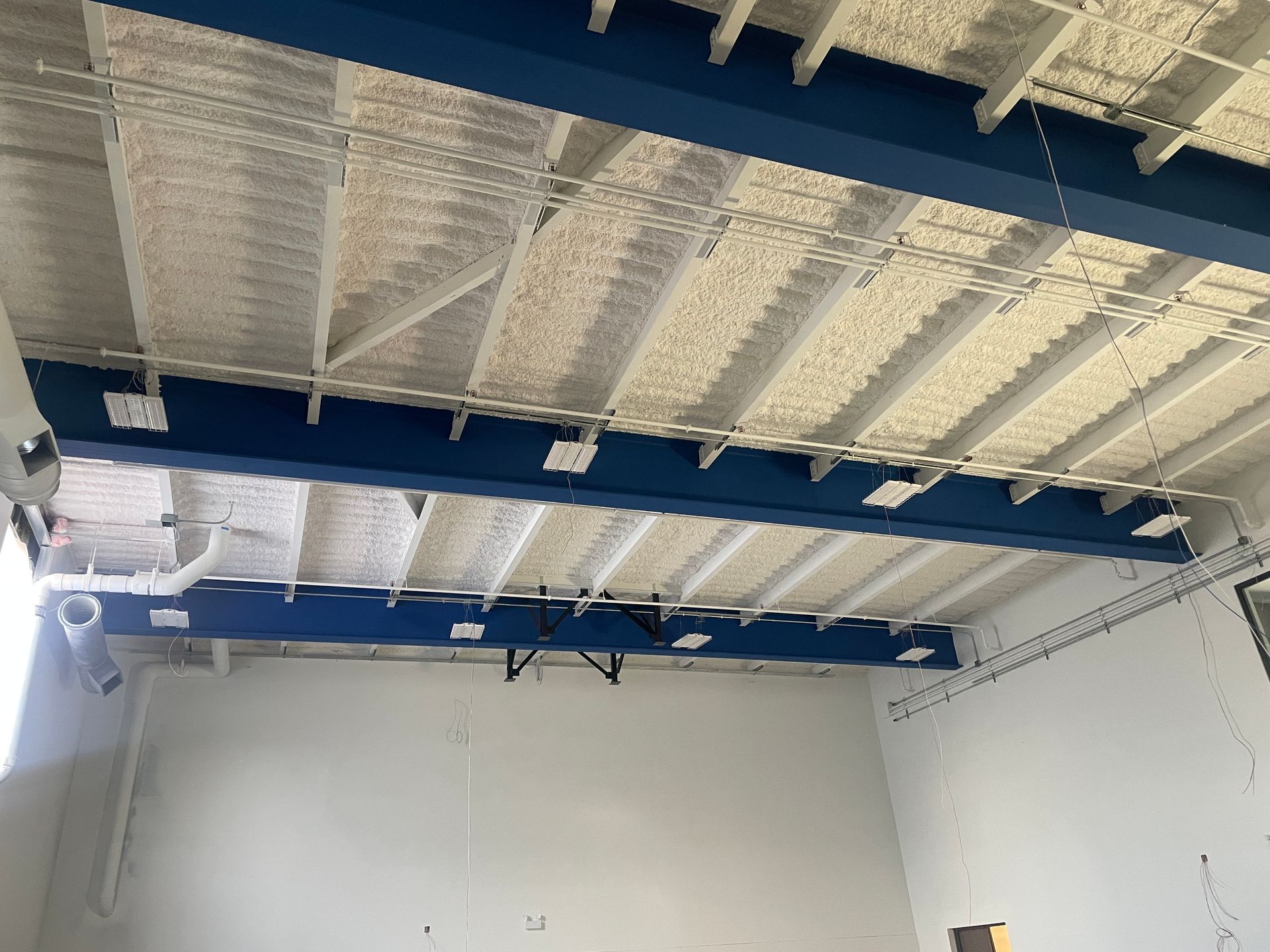 White room with a blue steel beam ceiling. Foam insulation is applied to the ceiling.