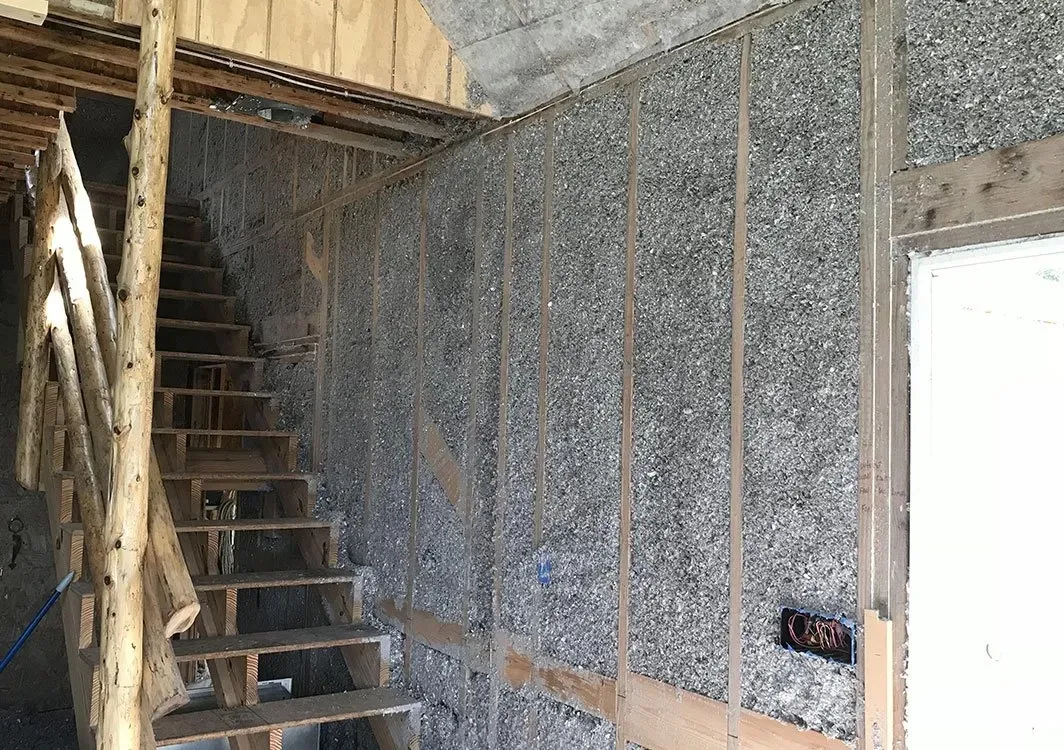 Interior view of a building under construction. Staircase and wall insulation are visible, with exposed wooden framing.