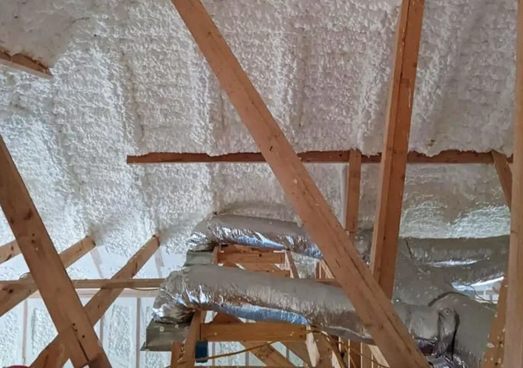 Interior view of attic insulation with spray foam between wooden beams and HVAC ductwork.