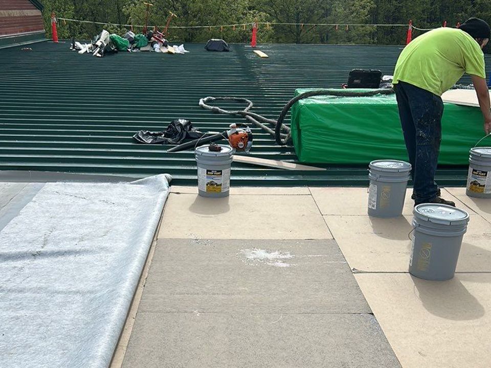 Person on a roof applying sealant with buckets and equipment, green metal roof in background.