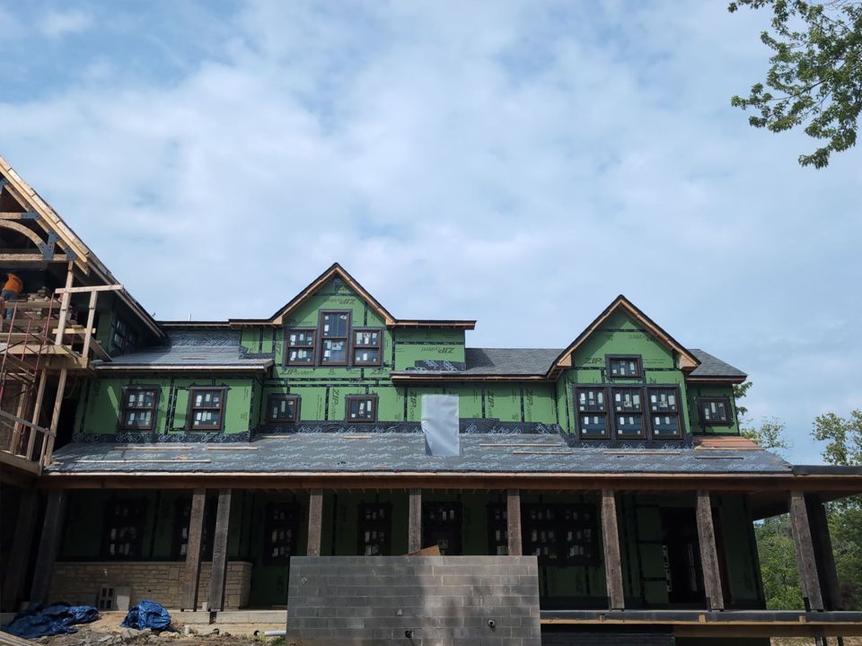 House under construction, green sheathing, dark windows, gray roof, wooden porch. Cloudy sky.