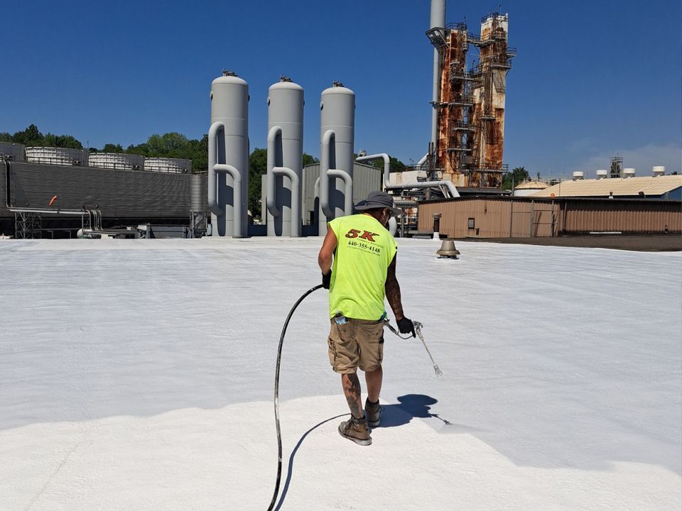 Person spraying white coating on a rooftop; industrial setting with tanks and a smokestack.