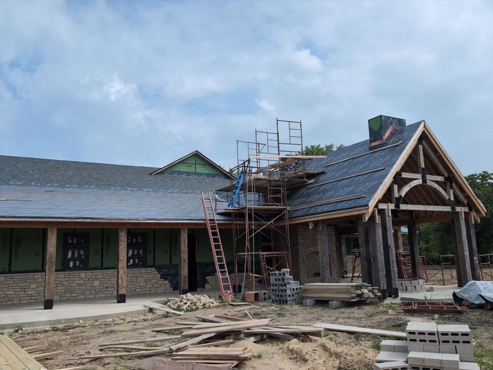 A single-story building under construction with a timber-frame porch, partial stone siding, and roof underlayment.
