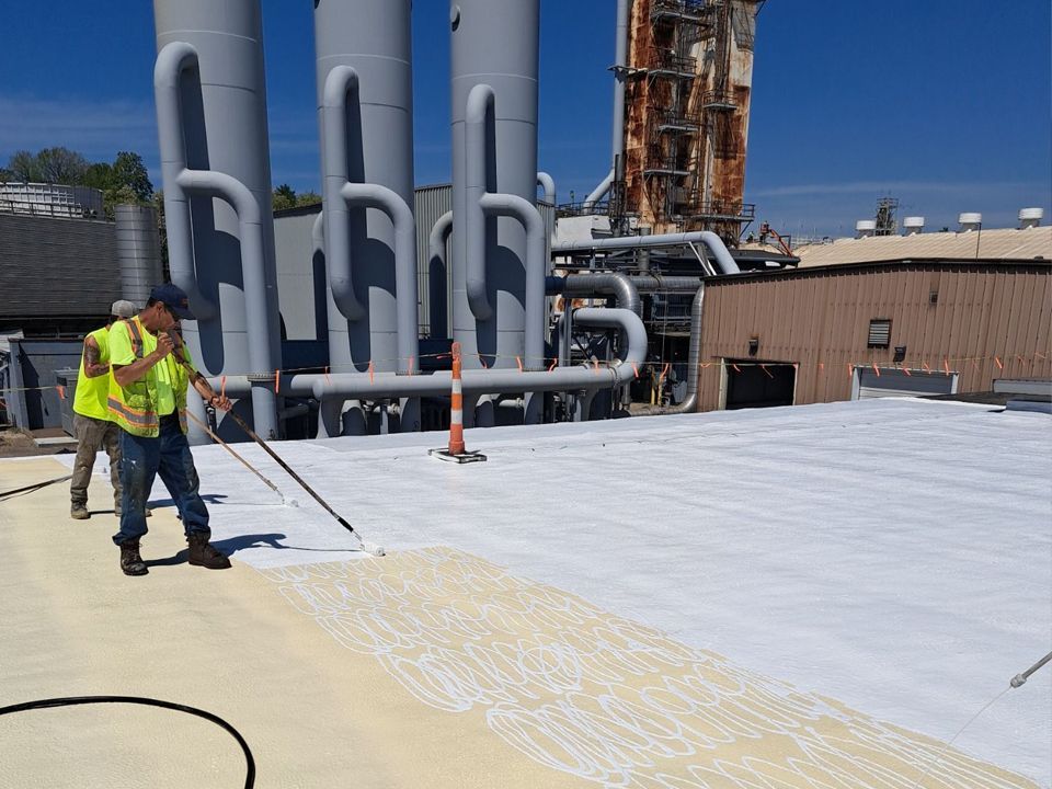 Workers applying white sealant to a roof near industrial equipment under a blue sky.