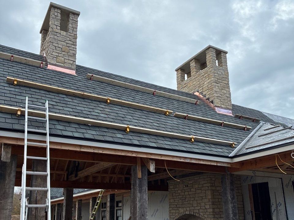 Roof with shingles and installed wood supports, chimneys, and a ladder under an overcast sky.