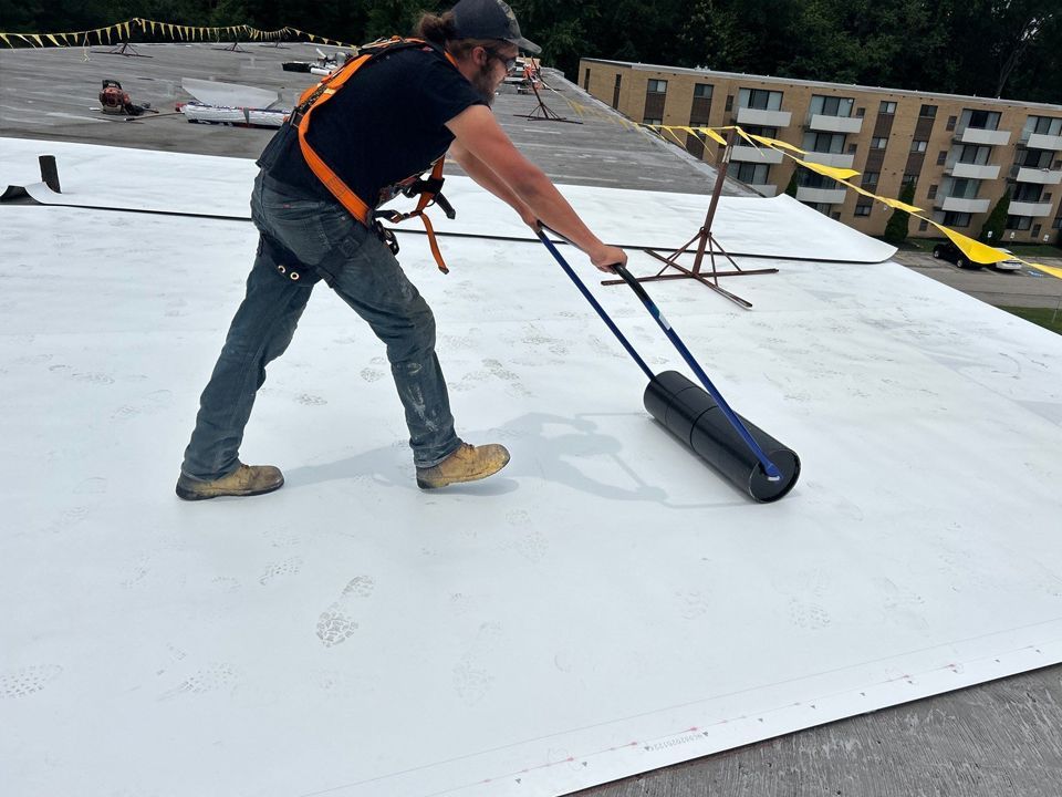 Person rolling roofing material on a flat roof, wearing safety harness.