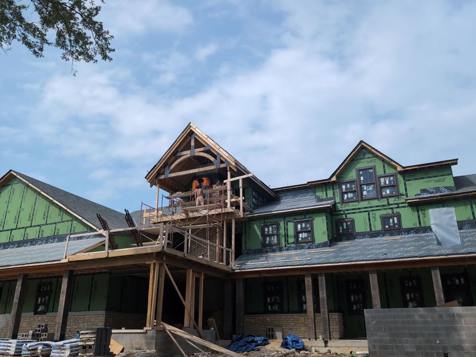 Construction of a large wooden house with multiple gables. Green wrap covers the frame, blue sky in background.