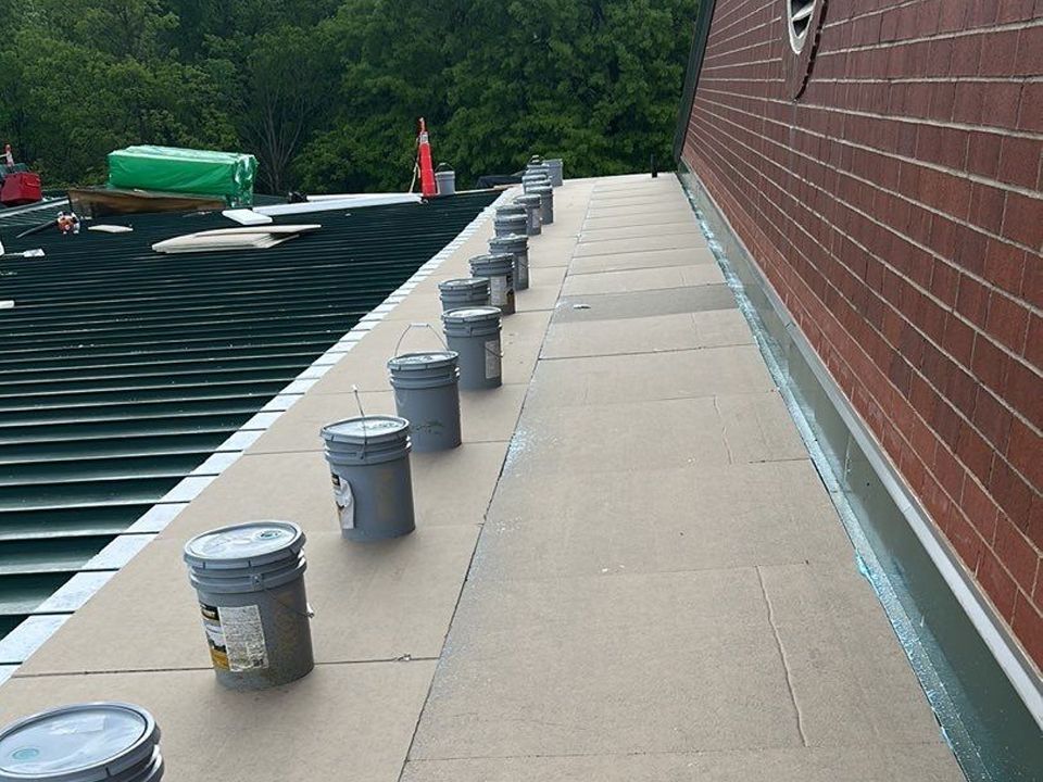Roofing work: buckets line a roof's edge next to a brick wall. Green tarp and trees are in the background.