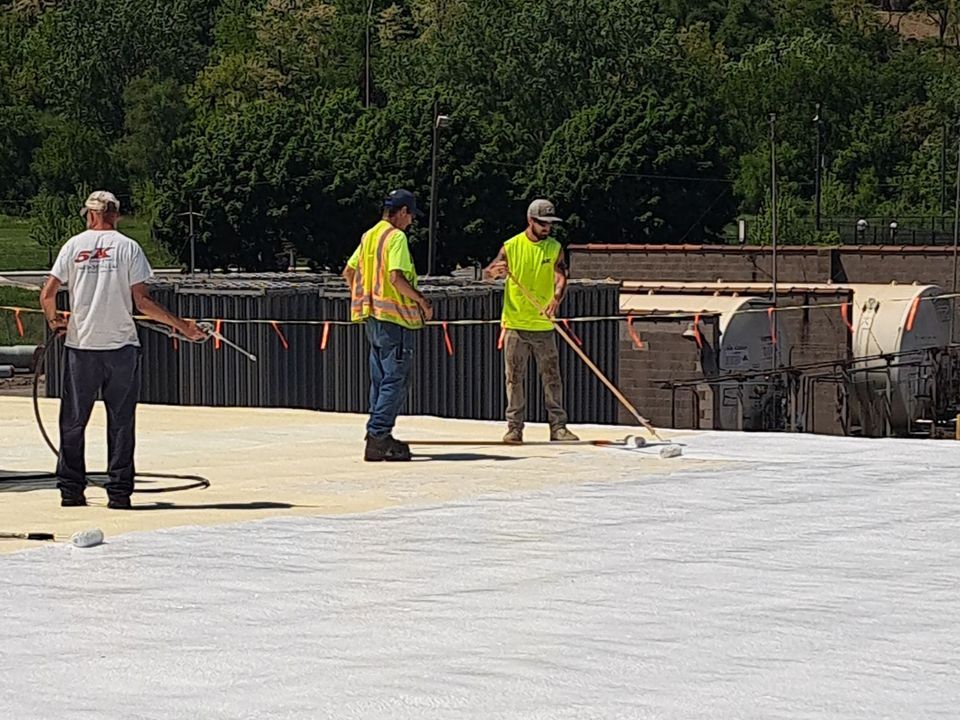 Three workers on a construction site applying a white sealant. Trees and concrete structures in the background.