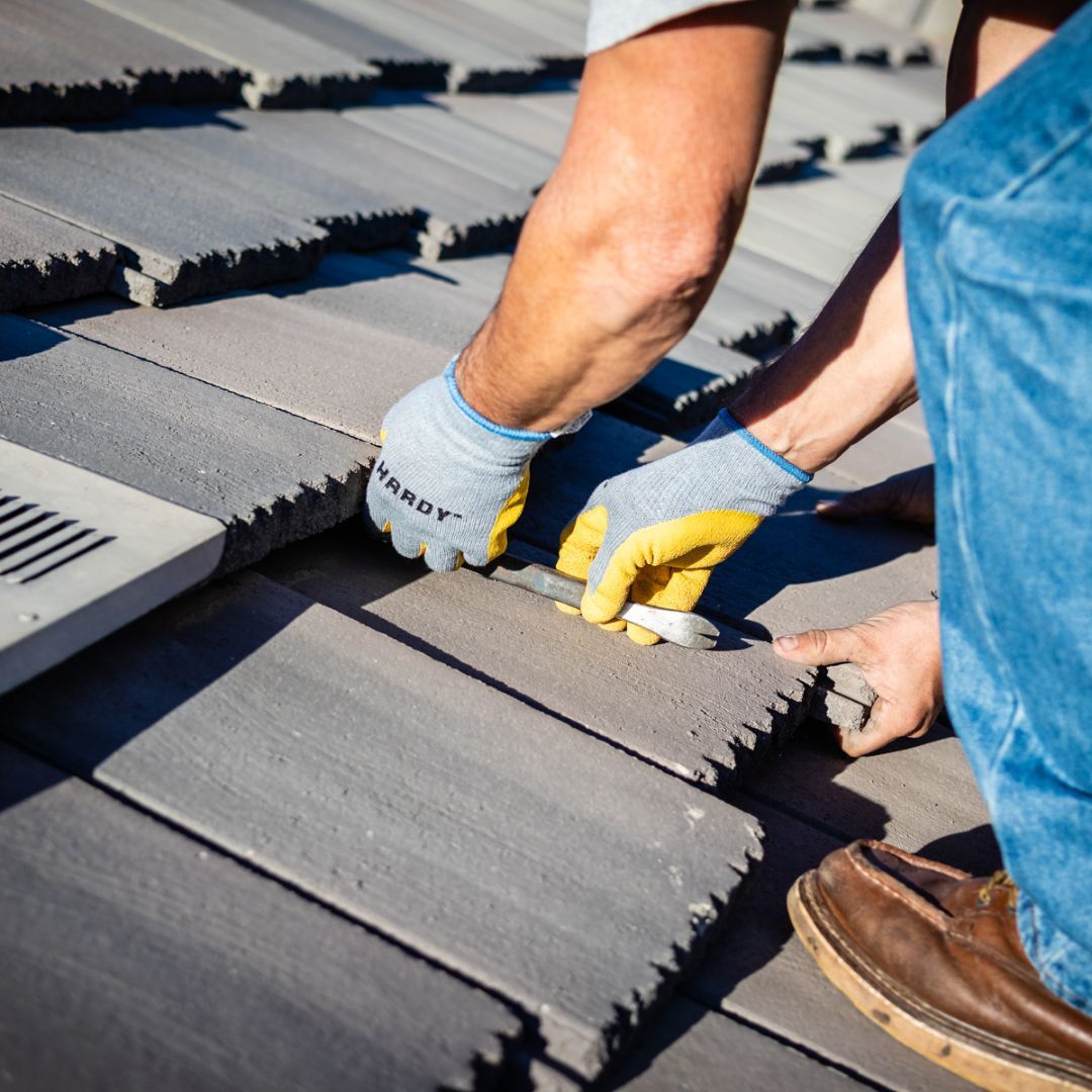 A person wearing work gloves repairs gray concrete roof tiles on a sunny day.