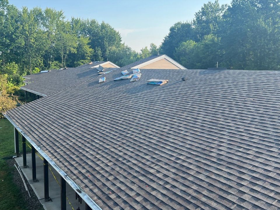 Gray shingle roof of a building, with multiple vents. Trees are visible in the background on a sunny day.