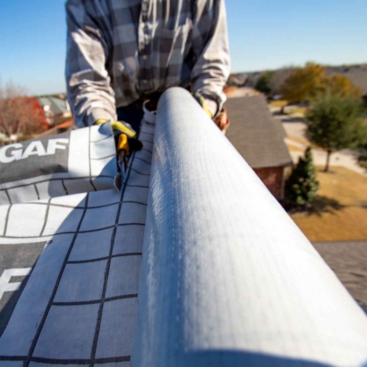 Person on a roof unrolling GAF roofing material with grid pattern. Autumn day, residential setting.