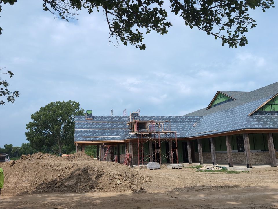 Building under construction with dark grey slate roof, scaffolding, and green trim. A pile of dirt in foreground.