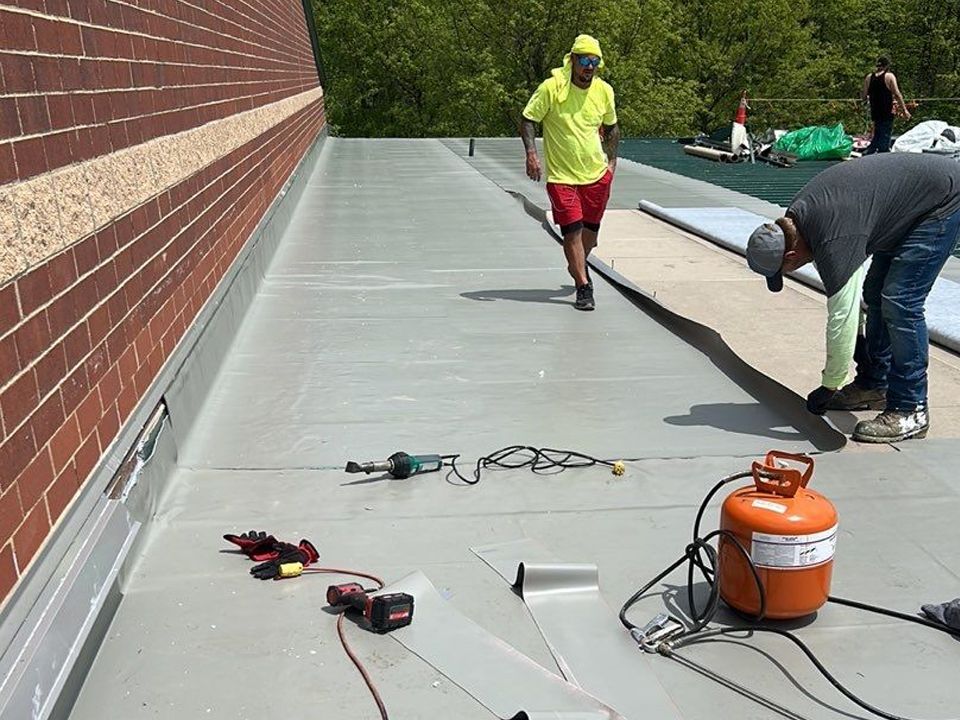 Workers installing roofing on a building with a brick wall. One worker walks, two others work near a propane tank.