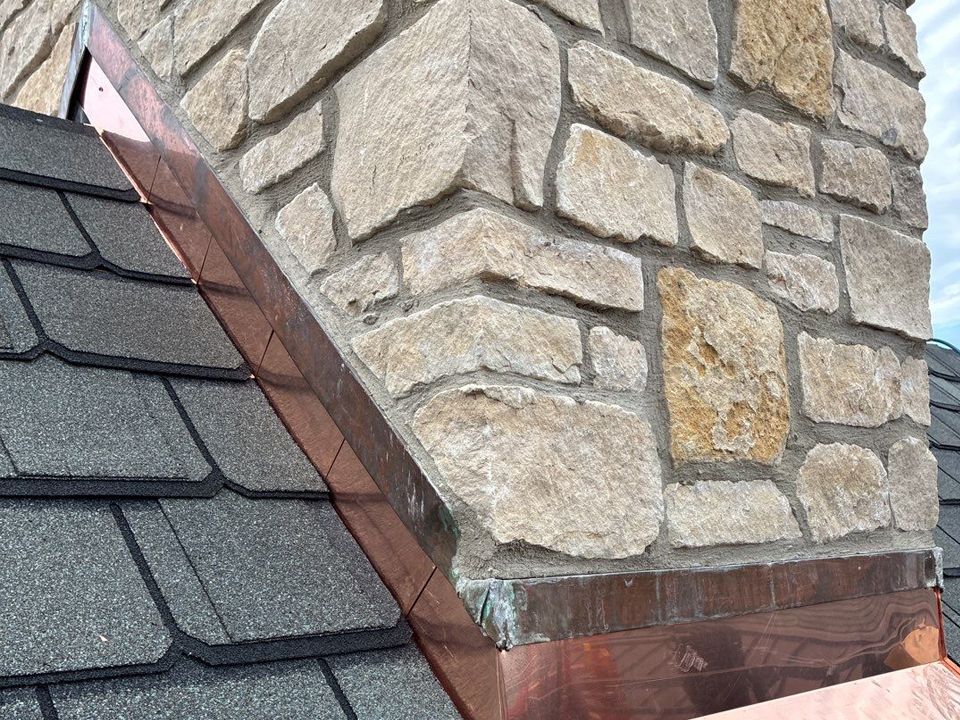 Copper flashing at the base of a stone chimney on a dark shingled roof.