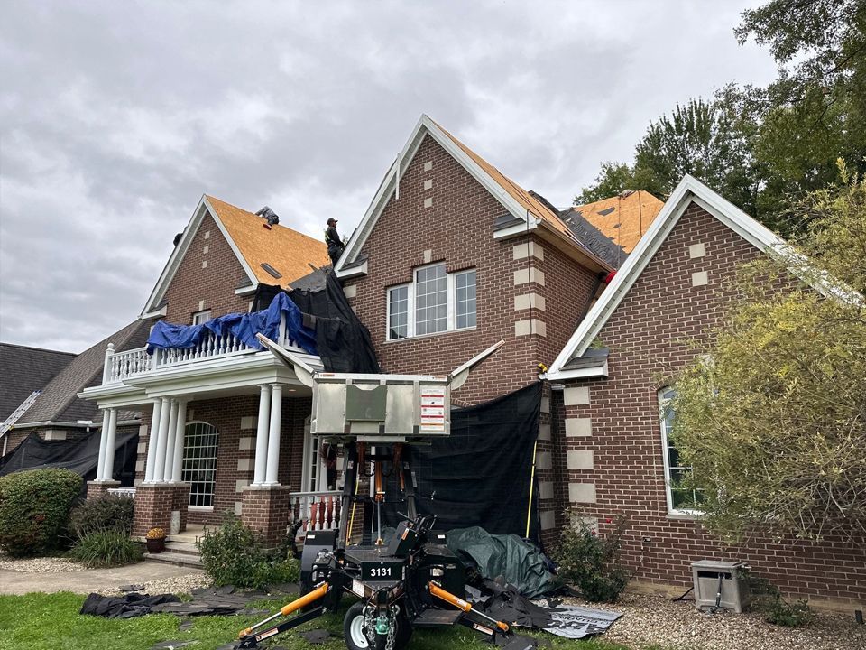 House with brick facade undergoing roof repair; black tarps and equipment visible.