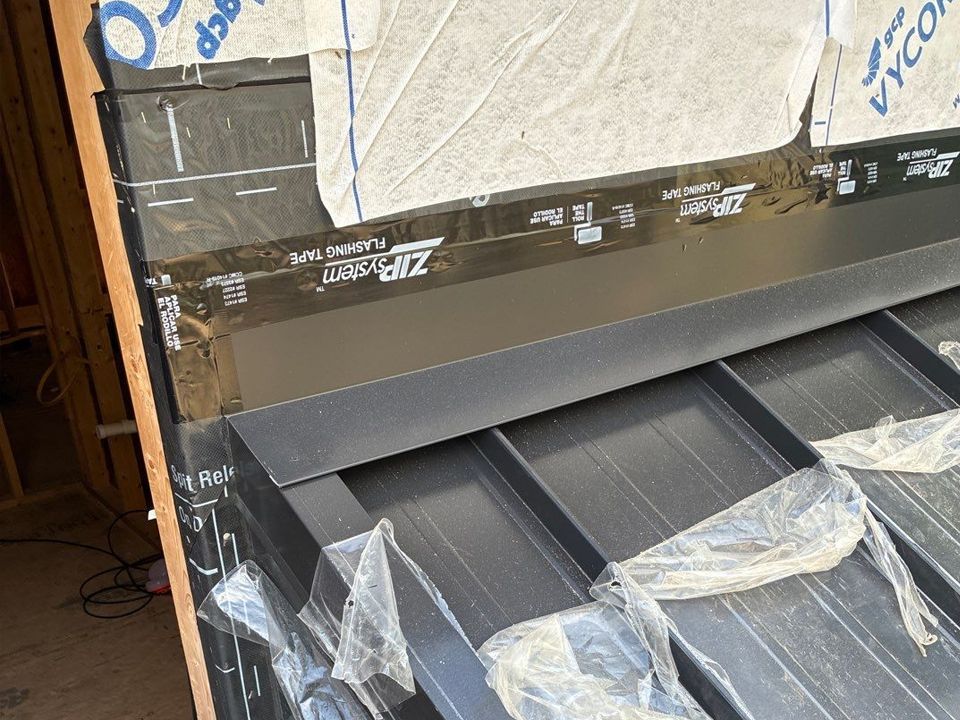 Construction of a metal roof with black siding and flashing against a wall covered in protective wrap.