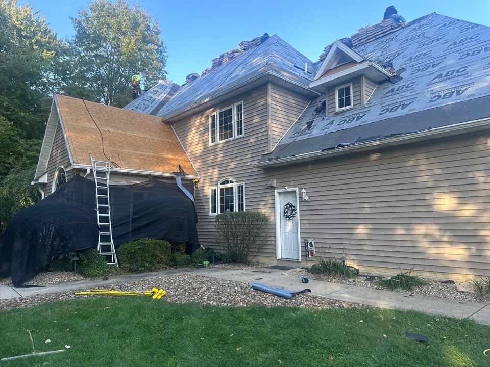 House roof partially covered with new shingles, construction in progress. Ladder and tarp visible.