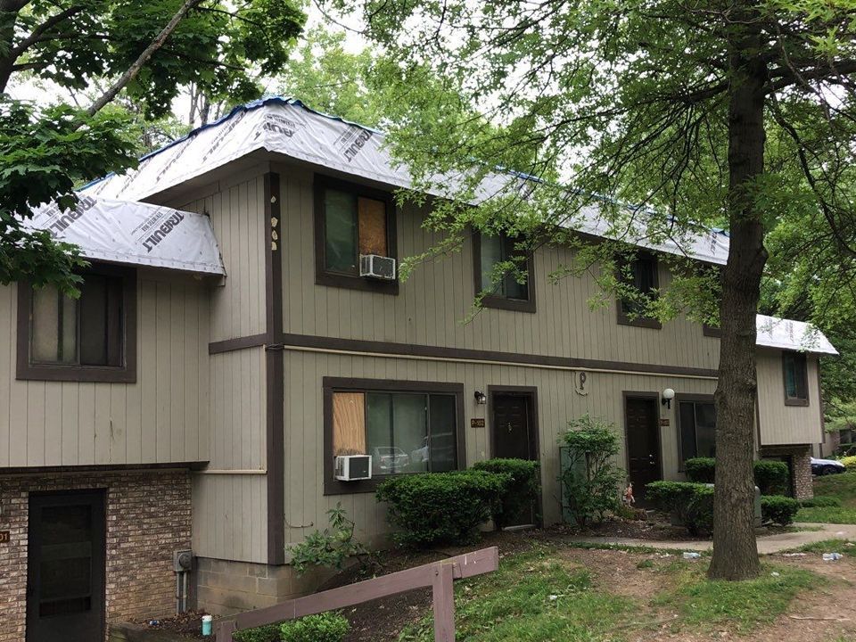 Two-story apartment building with tan siding, brown trim, and a partially covered blue roof, surrounded by trees.