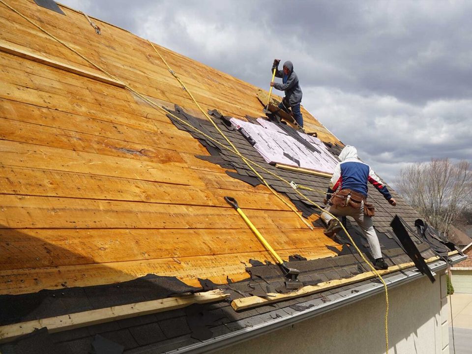 Two workers repairing a roof, removing old shingles. One uses a tool, secured by ropes. Cloudy sky.