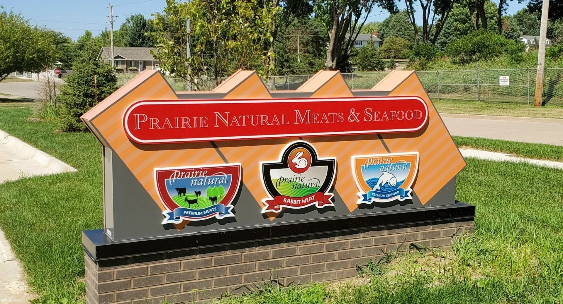 Sign for Prairie Natural Meals & Spreads with multiple logos on a brick base and grass surroundings.