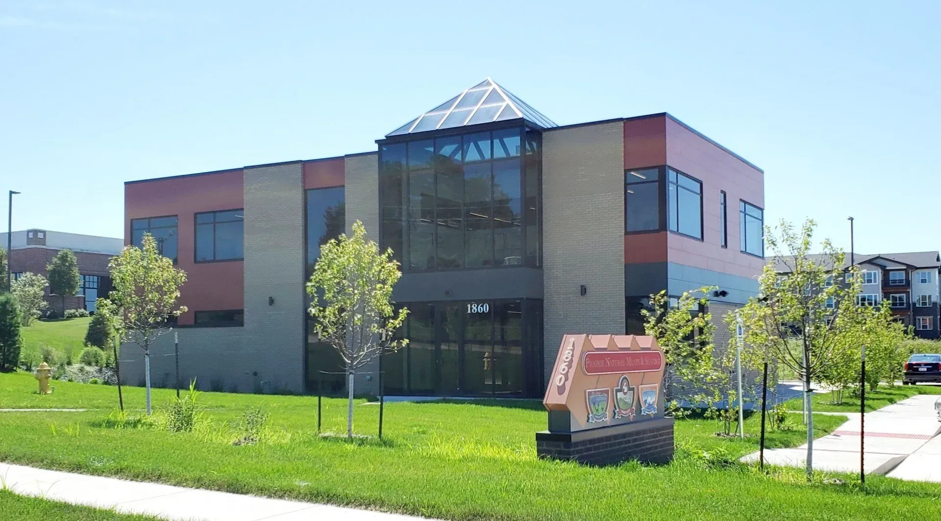 Two-story brick and glass building with trees and signage on a grassy lawn under a blue sky.