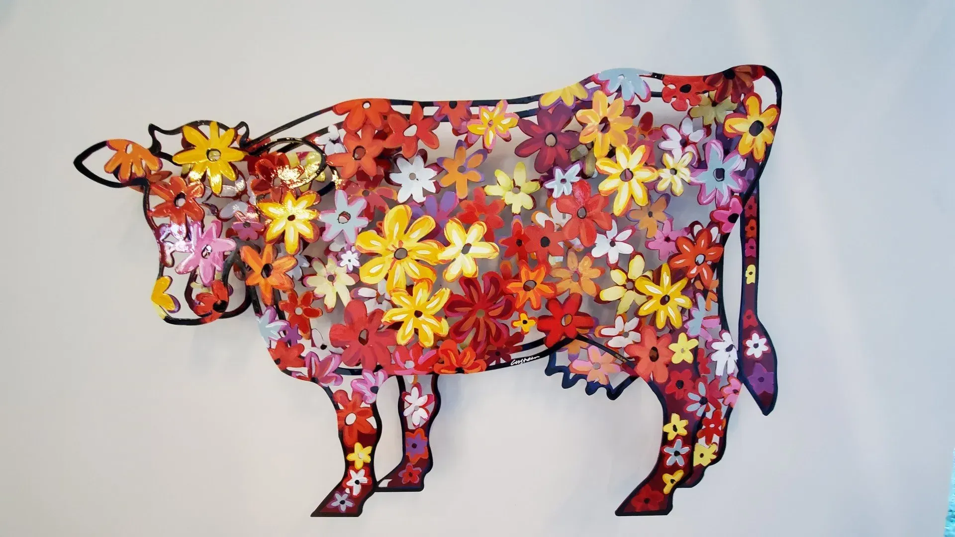 Floral cow art. Black outline filled with colorful paper flowers against a white wall.