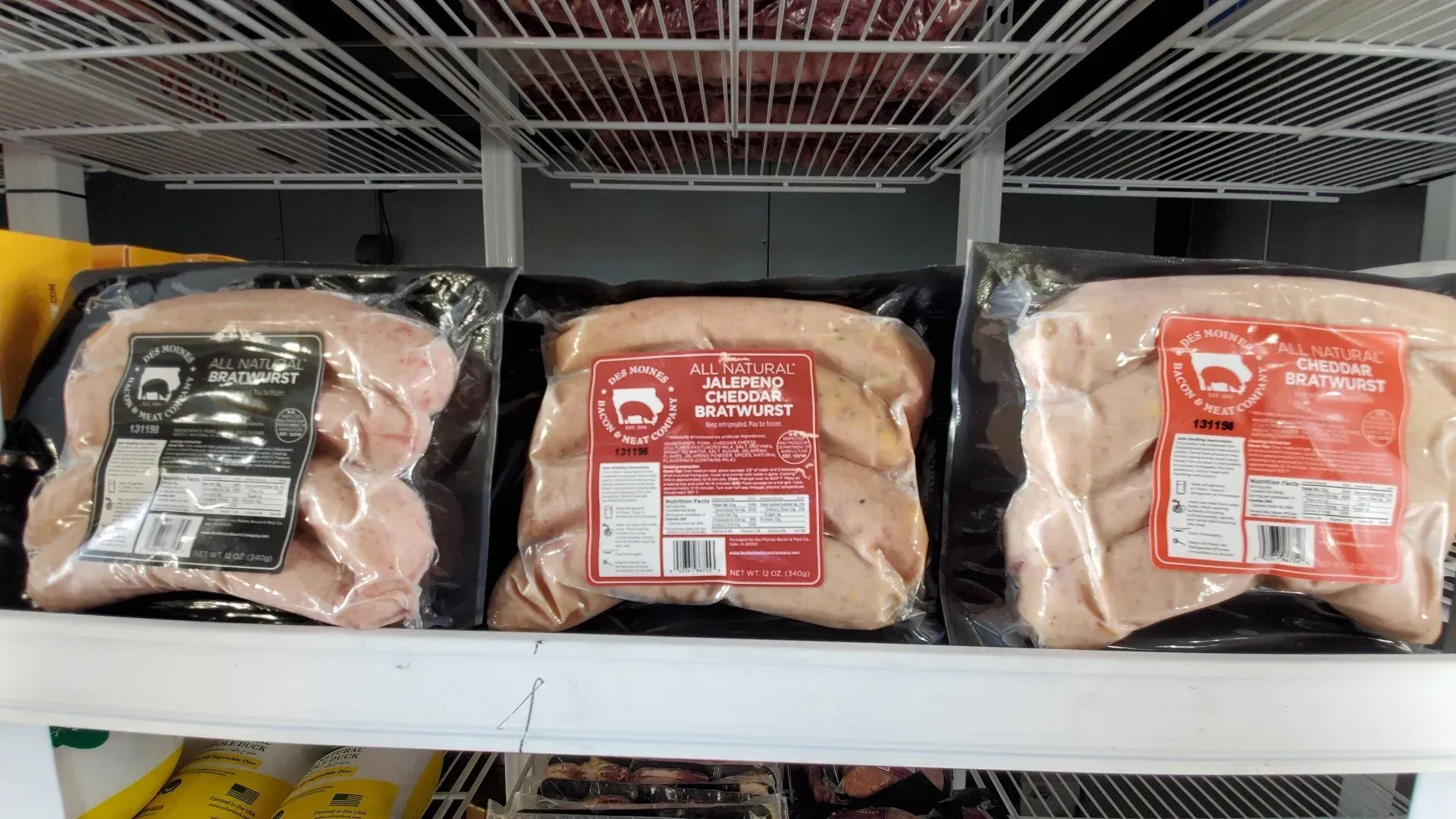 Three packages of raw meat sausages on a refrigerated shelf, under white wire shelving.