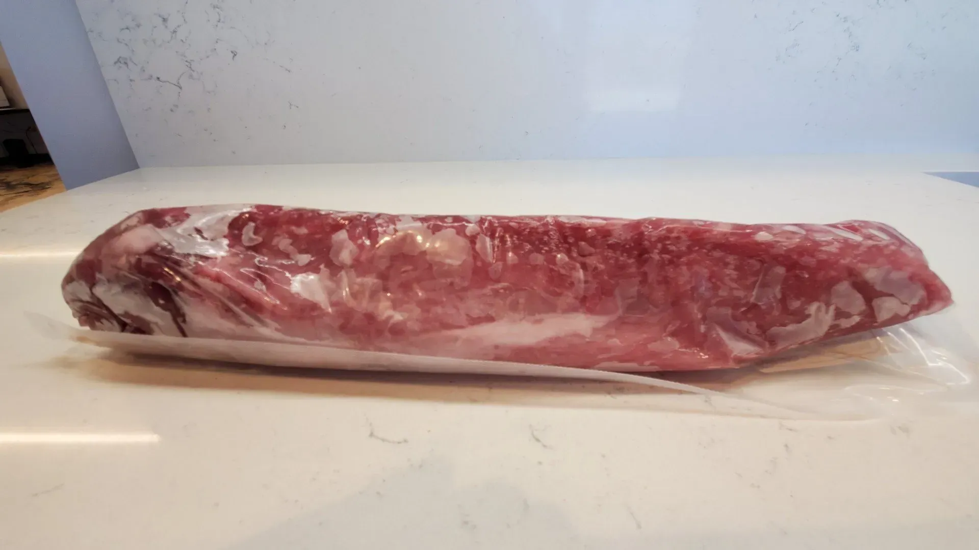 Raw pork tenderloin in plastic wrap on a white surface.