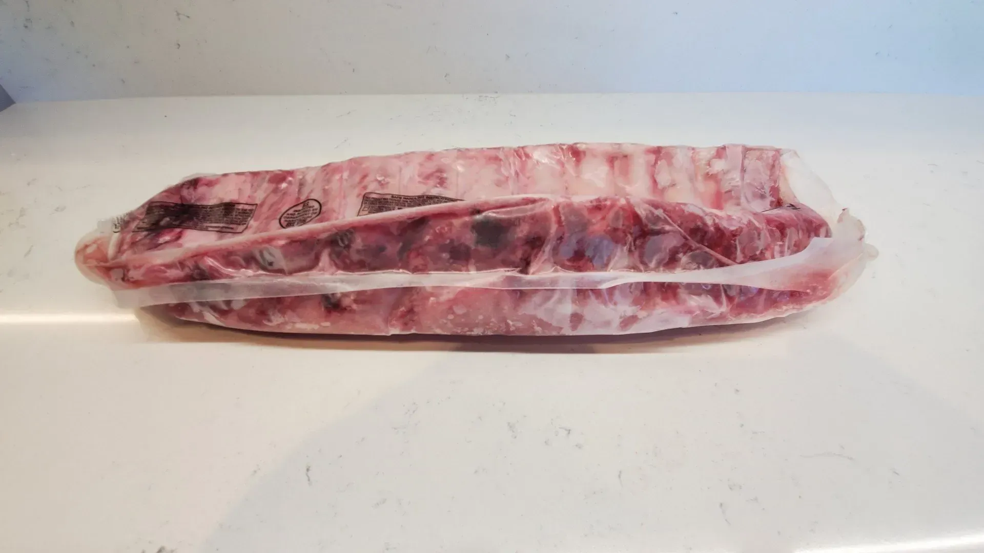 Rack of raw ribs sealed in plastic wrap on a white surface.