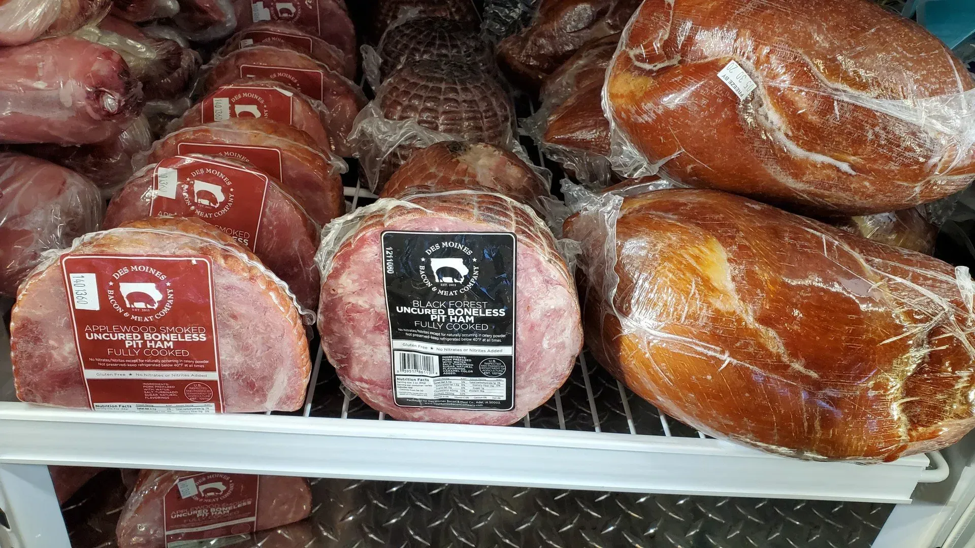 Hams on a shelf, wrapped in plastic. Some have labels with a cow graphic, in a cooler.
