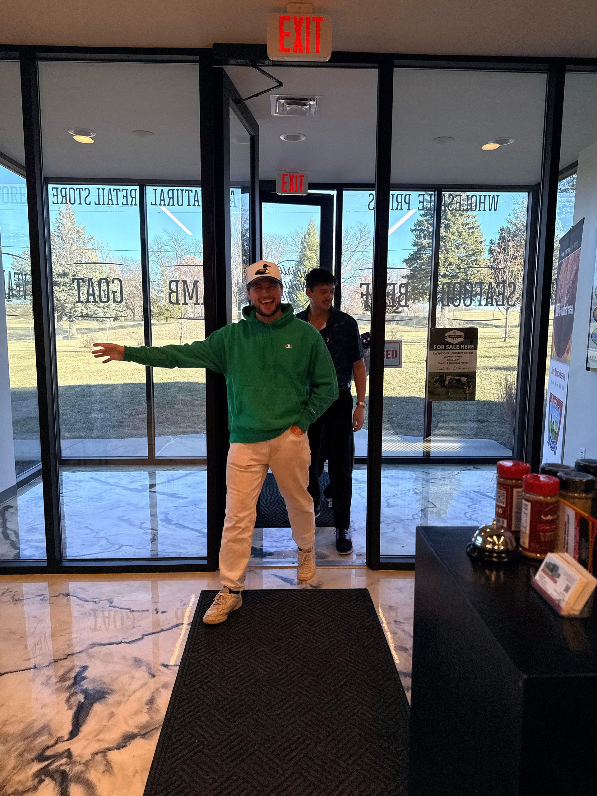 Person in green shirt and beige pants stands in a glass entrance doorway of Customers walking up to the entrance of Prairie Natural Meats & Seafood.
