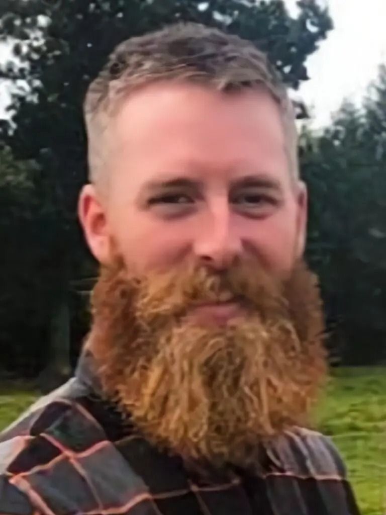 A man with a beard is wearing a plaid shirt and smiling.