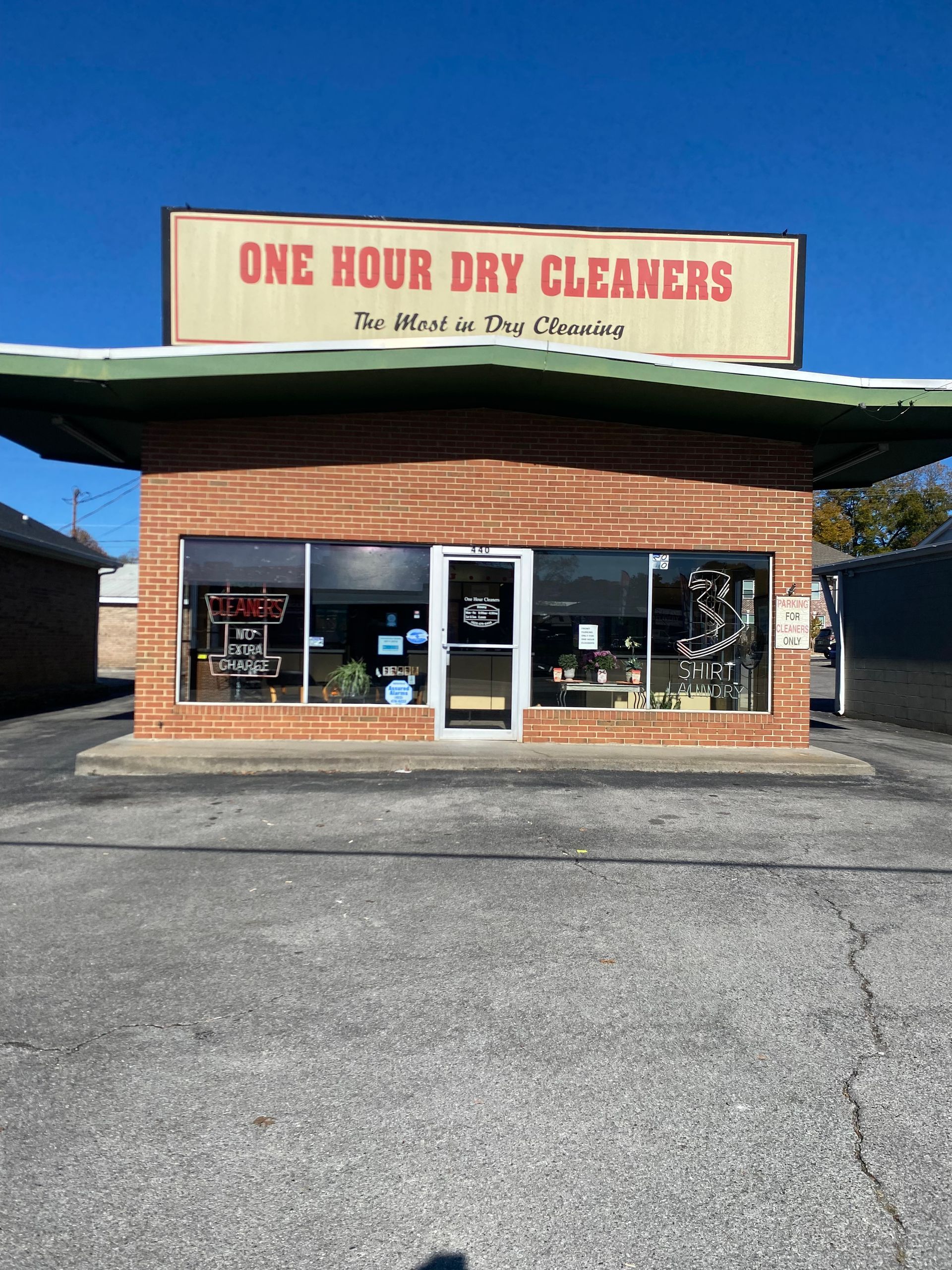 One Hour Dry Cleaners building with red brick, glass windows, and a sign. Blue sky and paved parking lot.