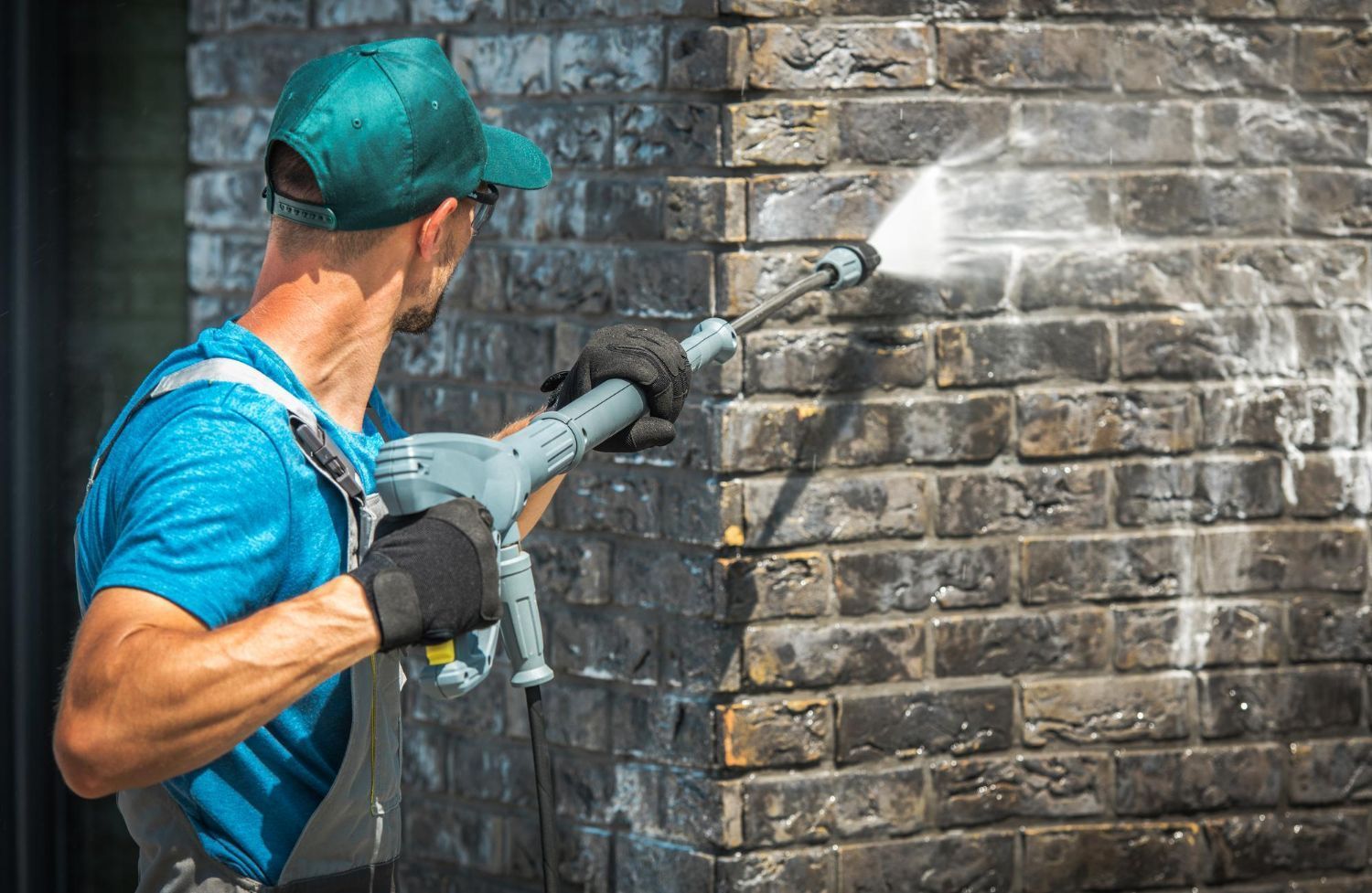 A person in a blue shirt and green cap uses a power washer to clean a dark brick wall.