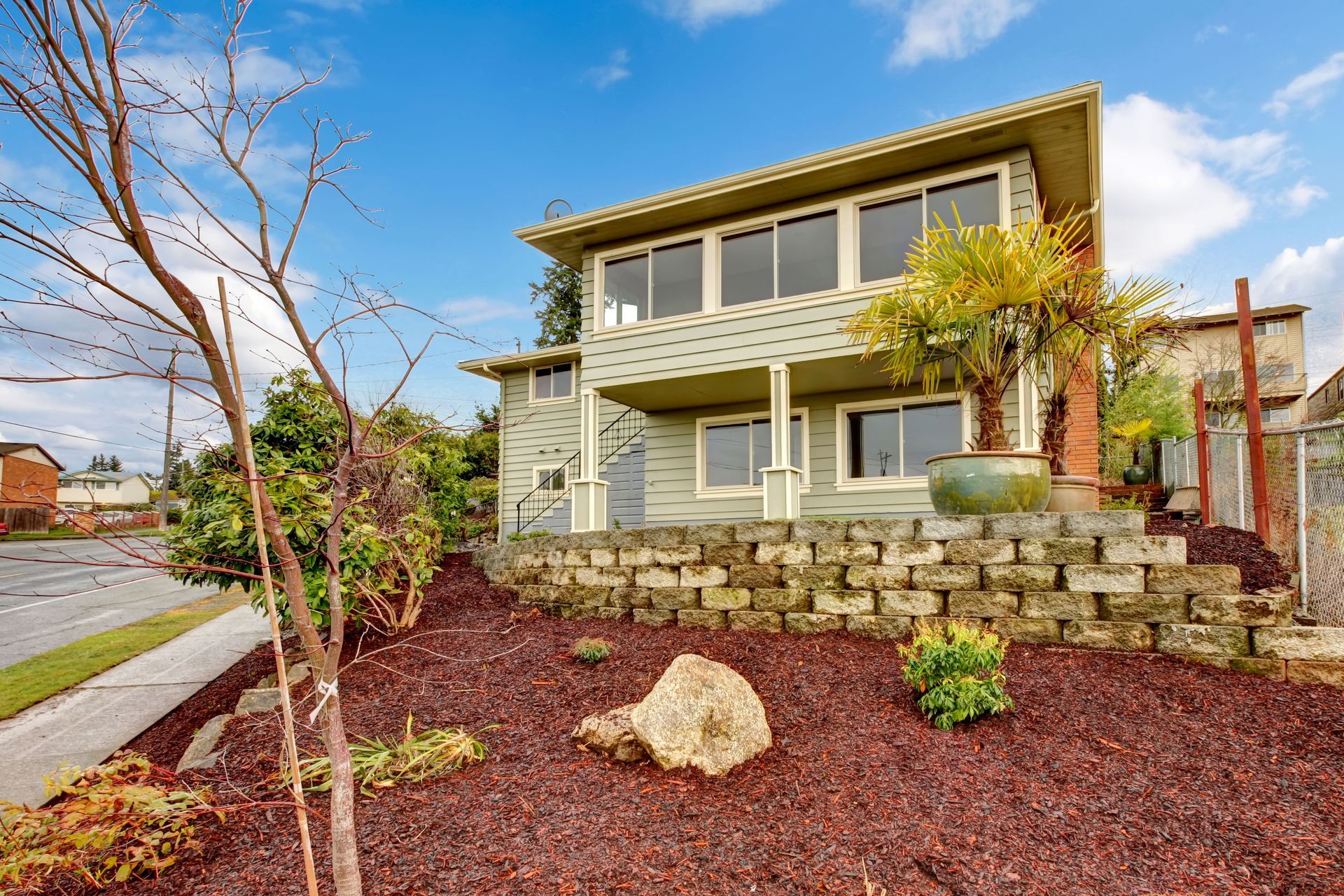 A light green two-story house with a terraced stone wall, a front yard with red mulch, and a potted palm tree.