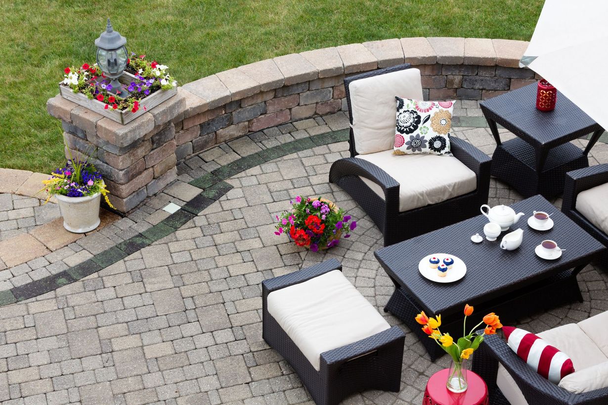An outdoor patio featuring wicker furniture, a table set for tea, potted flowers, and a curved stone retaining wall.