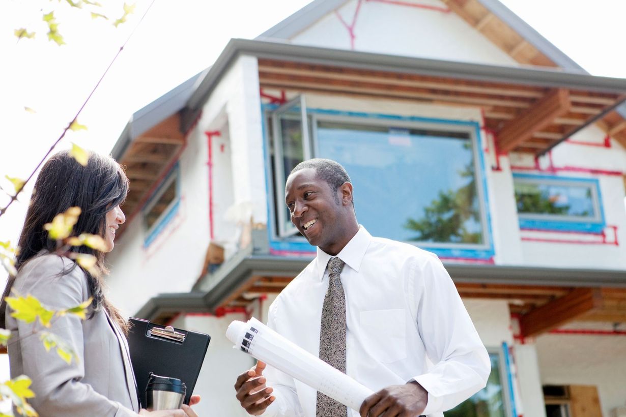 A professional man holding blueprints talks to a colleague in front of a modern house under construction.